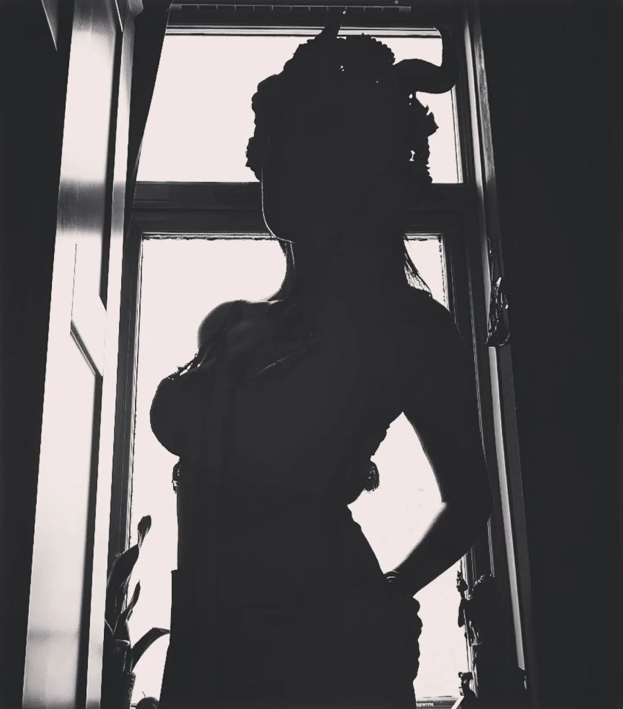 A black and white silhouette picture of Gemma. She's stood in front of a bright window wearing a horned headdress.